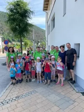 Eine Gruppe von Menschen, darunter Erwachsene und Kinder, steht zusammen auf einem gepflasterten Bürgersteig. Sie tragen Rucksäcke und Hüte, einige Kinder halten Schilder. Hinter ihnen steht ein Gebäude, Bäume und Berge.