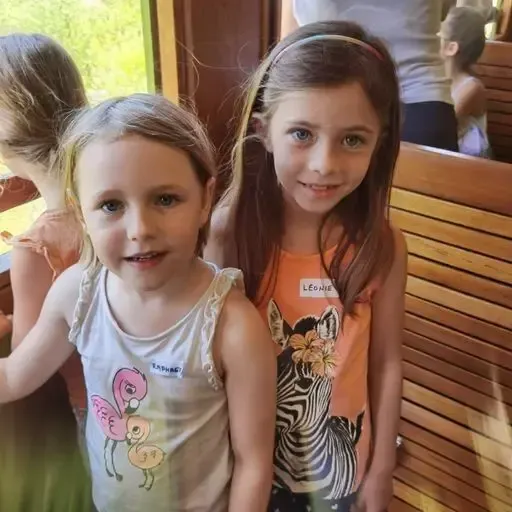 Zwei Mädchen in ärmellosen Shirts lächeln für die Kamera. Eine trägt ein Zebra, die andere einen Flamingo. Sie stehen auf einer Holzbank mit anderen Kindern im Hintergrund.