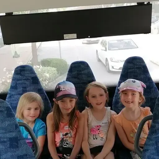 Vier junge Mädchen sitzen in einem Bus mit blauen Sitzen. Sie lächeln in die Kamera. Draußen auf der Straße fährt ein weißer Wagen vorbei.