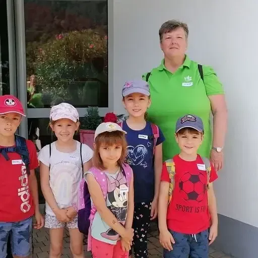 Eine Frau und fünf Kinder stehen vor einem Gebäude. Die Frau trägt ein grünes Shirt, und die Kinder haben Mützen und Rucksäcke an. Dahinter befindet sich ein Glasfenster mit Pflanzen und Blumen.