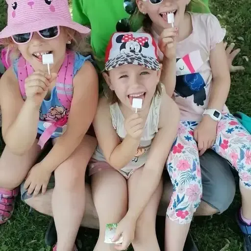 Drei junge Mädchen sitzen auf dem Gras. Sie lächeln und halten Eislutscher in der Hand. Eine trägt einen rosa Hut und eine Sonnenbrille, eine andere einen Mickey-Mouse-Hut und die dritte eine Sonnenbrille.