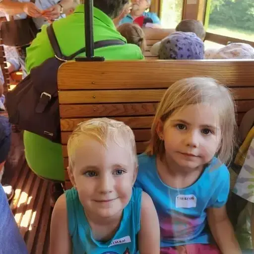 Zwei Kinder mit Namensschildern sitzen auf einer Holzbank, wahrscheinlich in einer Straßenbahn oder einem Zug, mit Erwachsenen und anderen Kindern im Hintergrund.