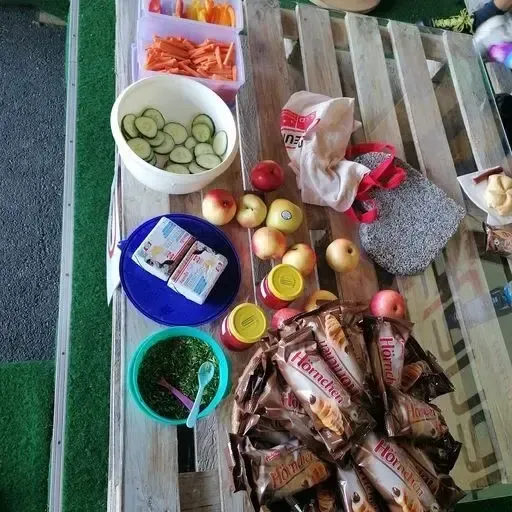 Ein Holztisch zeigt eine Schüssel mit geschnittenen Gurken, Äpfeln, verschiedene Snacks und einige Lebensmittel auf einem blauen Teller, zusammen mit einer Handtasche und einem Glas.