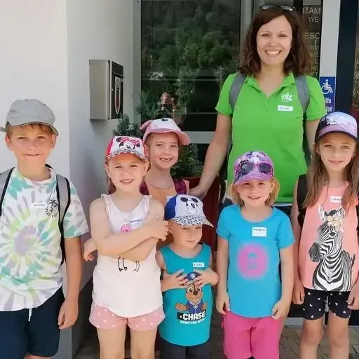 Eine Gruppe von Kindern und einer Frau stehen vor einem Gebäude. Die Frau lächelt und die Kinder tragen Hüte. Einige der Kinder halten eine kleine Pflanze.