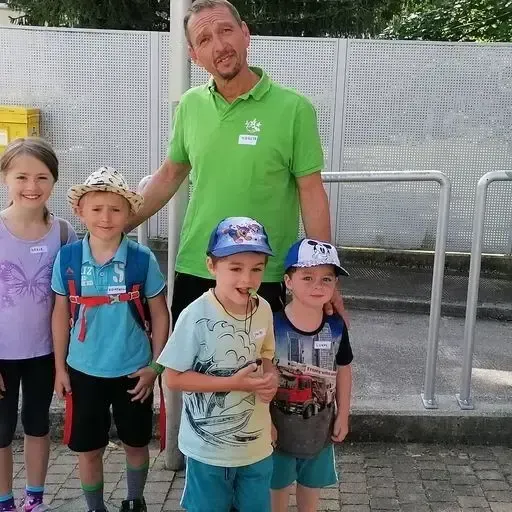 Ein Mann in einem grünen Poloshirt steht mit drei Kindern, zwei Jungen und einem Mädchen, in der Nähe eines Metallzauns auf einem gepflasterten Bürgersteig.