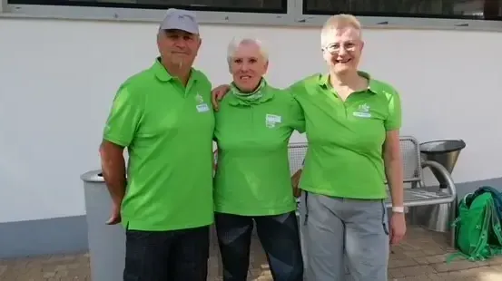 Drei Personen in grünen Shirts stehen nebeneinander und lächeln. Ein Mann links, eine Frau in der Mitte und eine Frau rechts. Sie stehen vor einer weißen Wand mit einer Bank dahinter.