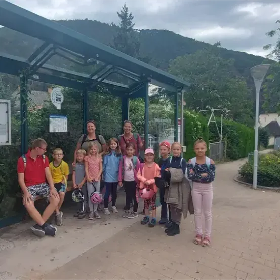 Eine Gruppe von Menschen steht vor einer Bushaltestelle, einige tragen Helme, mit Bergen und einem Haus im Hintergrund.