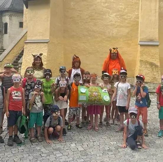 Eine Gruppe von Kindern und Erwachsenen posiert für ein Foto, alle tragen Tiermasken. Sie stehen vor einem Gebäude mit einer Treppe.