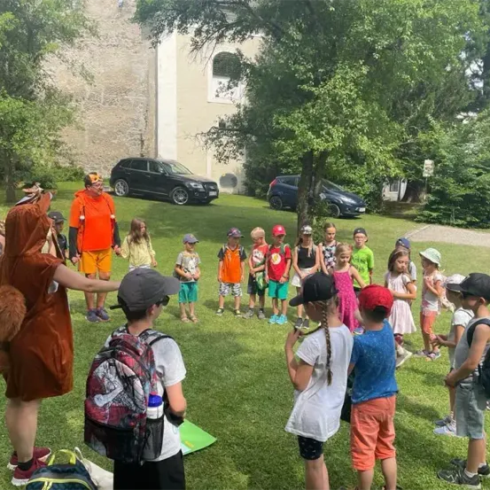 Eine Gruppe von Kindern in einem Park mit einem verkleideten Erwachsenen, zwei geparkten Autos und einem Gebäude mit einem Fenster im Hintergrund.