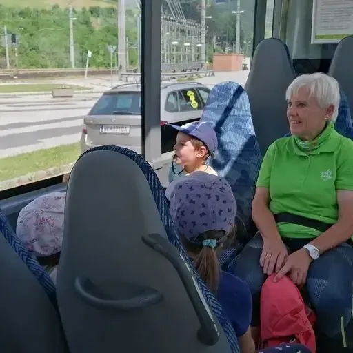 Eine ältere Frau und ein Kind sitzen auf einem Bus und sehen sich an, mit einem dritten Kind dahinter. Der Bus ist draußen geparkt, mit einem Auto in der Nähe.