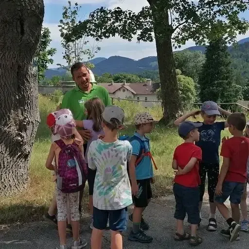Eine Gruppe von Kindern, einige mit Mützen, steht um einen Mann in einem grünen Hemd in der Nähe eines Baumes. Sie sind auf einem Schotterweg mit einem Grasbereich und Bergen im Hintergrund.