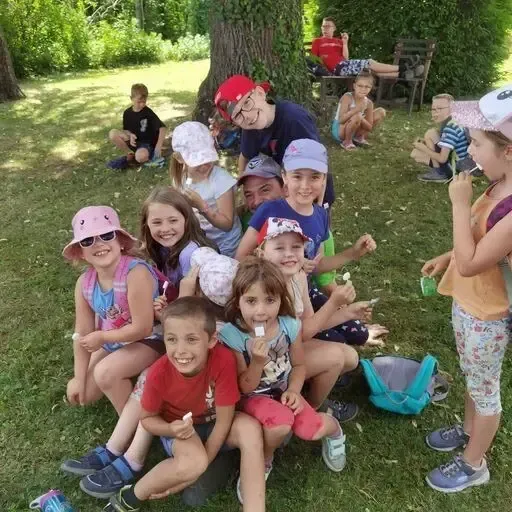 Eine Gruppe von Kindern und Erwachsenen sitzt auf dem Rasen, einige essen Eis. Einige tragen Hüte und Brillen. Ein Mädchen hält einen Lollipop. Ein Mann sitzt auf einer Bank hinter ihnen.