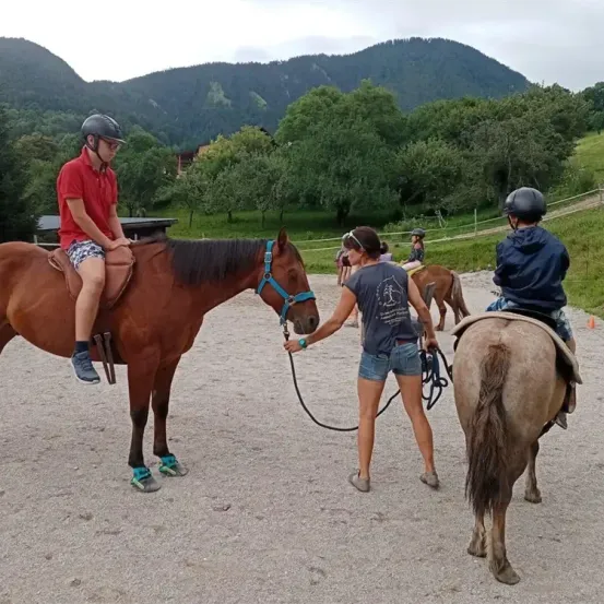 Eine Gruppe von Menschen reitet Pferde in einem Freiluftbereich. Ein Mann hält die Zügel des Pferdes, auf dem er reitet. Eine andere Person reitet ein Pferd und hält ein Seil. In der Ferne gibt es Berge und Bäume.