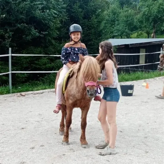 Ein Mädchen reitet ein kleines braunes Pferd, während ein anderes Mädchen die Zügel hält in einem eingezäunten Bereich. Bäume und ein Gebäude sind im Hintergrund.