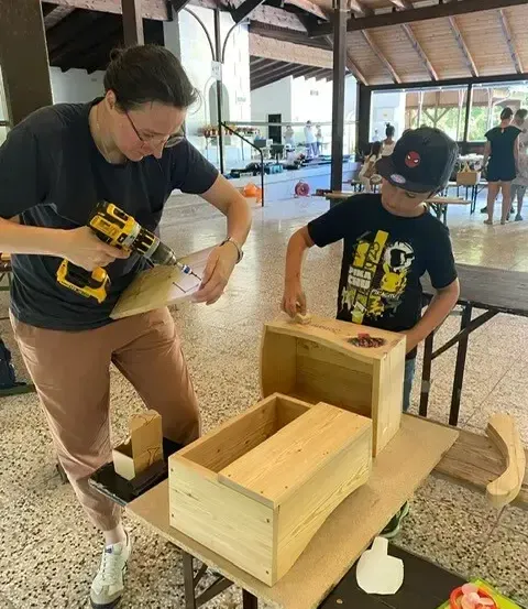 Eine Frau und ein Junge arbeiten an einer Holzkiste, mit einer Bohrmaschine und Schleifpapier. Die Szene ist drinnen, mit einer Holzdecke und einem gefliesten Boden. Andere Menschen sind im Hintergrund zu sehen.