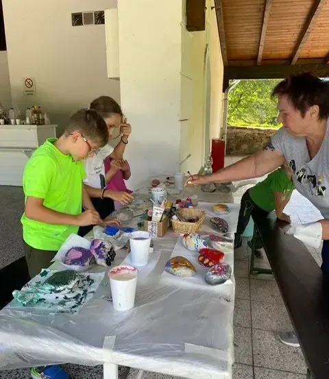 Eine Frau und zwei Kinder malen an einem Tisch mit Plastiktischdecke, bedeckt mit Tassen und Körben voller Farbe.