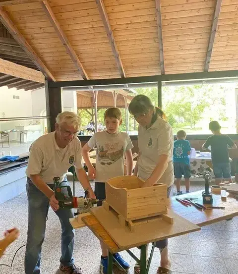 Ein älterer Mann und ein junger Junge arbeiten an einer Holzkiste, während eine Frau sie justiert. Hinter ihnen spielt ein Kind in der Nähe eines Tisches mit einem Laptop. Die Umgebung ist drinnen mit Holzbalken an der Decke.