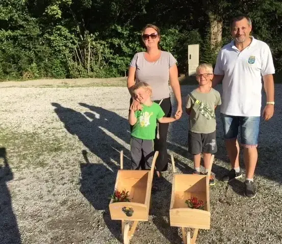 Eine vierköpfige Familie steht in einem Kiesbereich und lächelt für die Kamera. Zwei Kinder halten kleine Holzwagen mit Blumen.