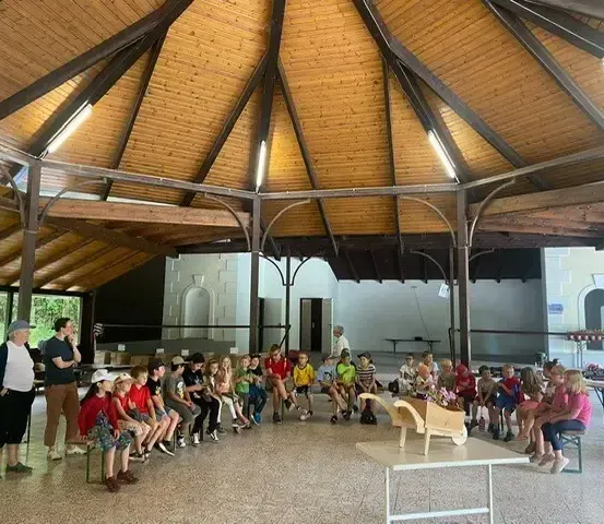 Eine Gruppe von Kindern und Erwachsenen sitzt in einem Kreis in einem Pavillon. Einige sitzen auf Stühlen, andere auf dem Boden. Eine Frau steht vorne und spricht möglicherweise. Ein Tisch mit einem hölzernen Schubkarren steht in der Mitte.