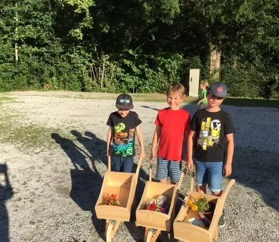 Drei Jungen stehen mit ihren hölzernen Schubkarren voller Blumen in einer Außenumgebung. Der Hintergrund zeigt Bäume und einen Kiesweg.