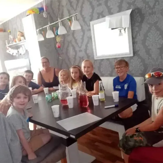 Eine Gruppe von Kindern und Erwachsenen sitzt an einem Tisch mit Getränken und Snacks. Der Tisch ist mit Bechern, Gläsern und Flaschen gedeckt. Der Raum hat eine tapezierte Wand und ein Fenster.