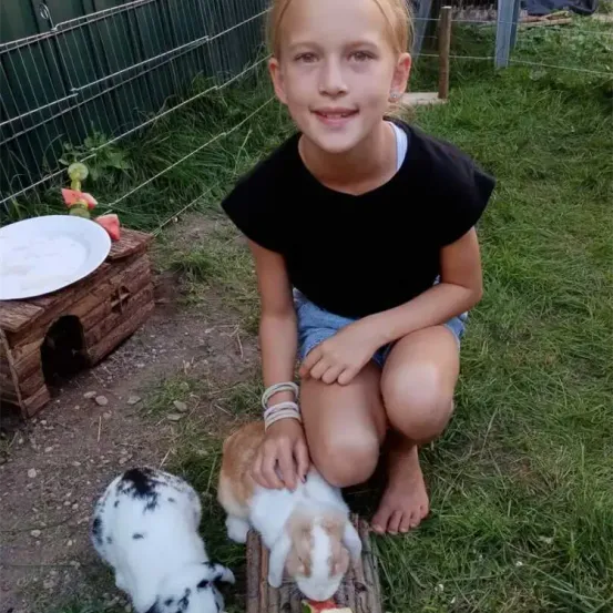 Ein junges Mädchen kniet lächelnd mit zwei Kaninchen im Garten. Sie hält ein Stück Futter für das Kaninchen auf der rechten Seite. Dahinter befindet sich ein Holzhaus mit einem Teller.