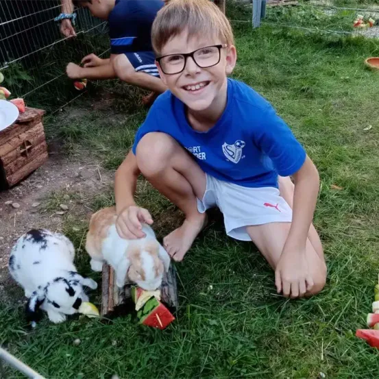 Ein junger Junge mit Brille und blauem T-Shirt füttert zwei Kaninchen mit Früchten auf einem Holzstamm. Ein weiterer Junge in einem blauen Shirt und Shorts sitzt hinter einem Zaun.