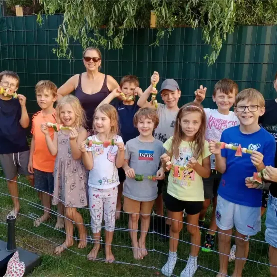 Eine Gruppe von Kindern und einer erwachsenen Frau steht in einer Reihe, lächelt und hält Essen. Sie scheinen sich bei einem Gartenfest zu befinden. Die Kinder halten verschiedene Essensartikel, und die Frau steht in der Mitte.
