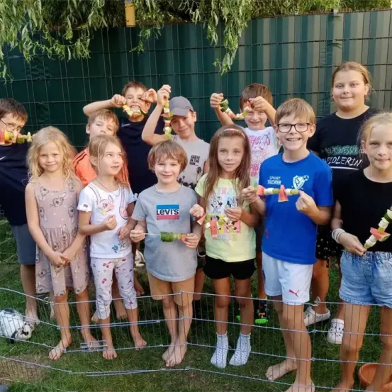 Eine Gruppe von Kindern steht in einem Garten mit einem grünen Zaun und hält Essen an Stöcken. Sie lächeln und scheinen für ein Foto zu posieren. Der Boden ist mit Gras bedeckt.