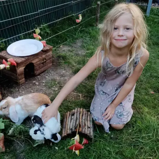 Ein kleines Mädchen mit blonden Haaren kniet im Gras und füttert zwei Kaninchen mit den Händen. Ein Holzhaus mit einem Teller darauf steht hinter ihr, und es gibt Wassermelonen an Stöcken.