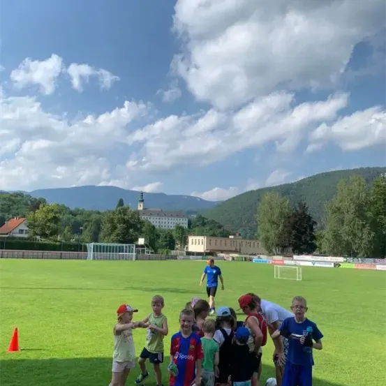 An einem sonnigen Tag versammeln sich eine Gruppe von Kindern und zwei Erwachsene auf einem Rasenfeld, vielleicht für ein Fußballtraining. In der Ferne befindet sich ein Gebäude mit Turm und Zaun.