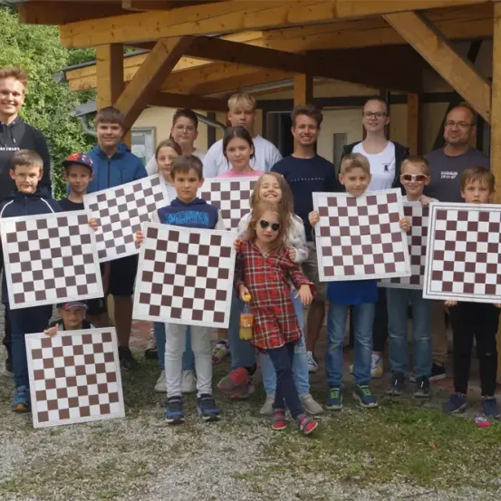 Eine Gruppe junger Kinder und Erwachsener steht vor einem Holzgebäude und hält große Schachbretter in der Hand, lächelt für ein Foto.