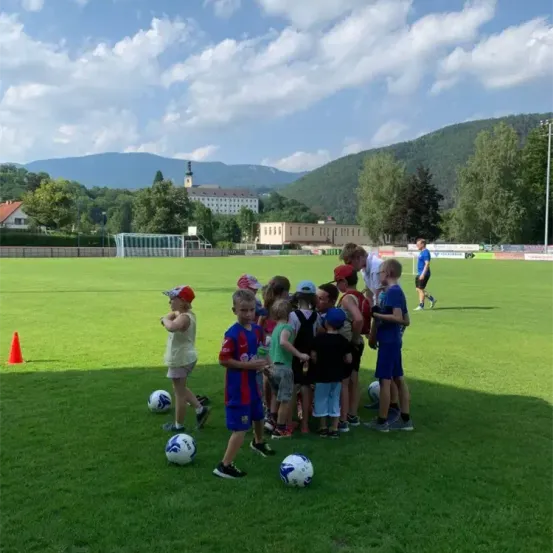 Kinder und Erwachsene versammeln sich auf einem Fußballfeld mit nahegelegenen Bällen. Im Hintergrund sind ein Gebäude und eine Bergkette zu sehen.