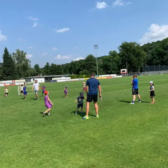 Eine Gruppe von Kindern und Erwachsenen spielt Fußball auf einem Feld unter einem teilweise bewölkten Himmel. Einige rennen, andere stehen, und es gibt einen Zaun und Bäume im Hintergrund.