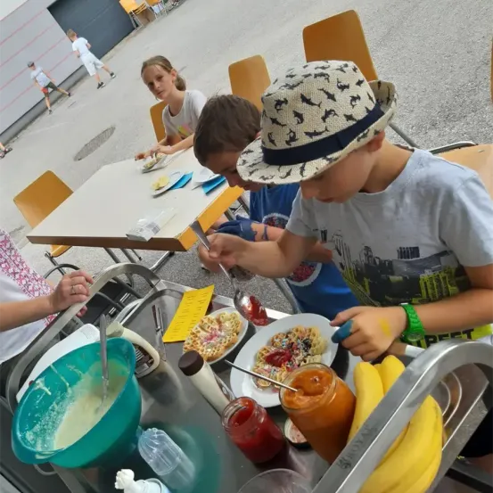 Kinder sitzen an einem Tisch und genießen eine Mahlzeit. Ein Kind fügt seinem Essen Sauce hinzu. Ein anderes Kind, das einen Hut trägt, greift nach Essen auf einem nahen Tisch. Es gibt verschiedene Gerichte, darunter Waffeln, Bananen und Beilagen.