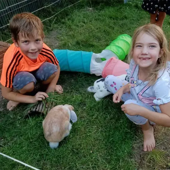 Zwei Kinder sitzen draußen mit einem Kaninchen. Der Junge spielt mit einem Kaninchenspielzeug, während das Mädchen zur Kamera schaut. Dahinter umschließt ein Zaun ein Kaninchengehege mit bunten Spielzeugen.