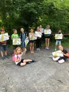 Eine Gruppe von Kindern steht und sitzt in einem Außenbereich und hält Karten hoch. Sie sind für Outdoor-Aktivitäten gekleidet. Der Hintergrund zeigt einen dichten Wald.