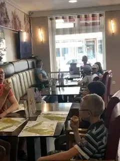 Ein Restaurant mit mehreren Gästen an Tischen. Ein Junge isst einen Snack, während eine Frau in der Nähe ein Menü liest. Der Raum hat Glasfenster und ist hell beleuchtet.