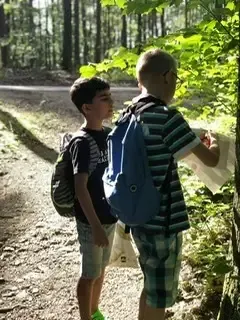 Zwei Jungen stehen auf einem Waldweg, einer hält eine Karte und der andere schaut sie an. Sie haben beide Rucksäcke.