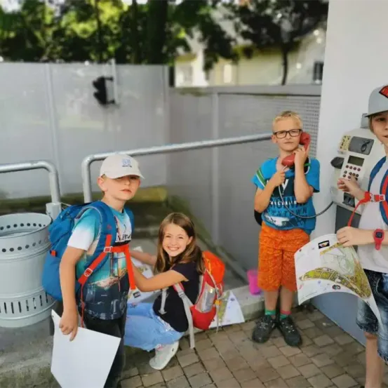 Vier Kinder stehen draußen, eines hält einen Stadtplan, ein anderes benutzt ein Telefon. Eines trägt einen Rucksack, und ein anderes hält ein Stück Papier. Ein Mülleimer ist in der Nähe.