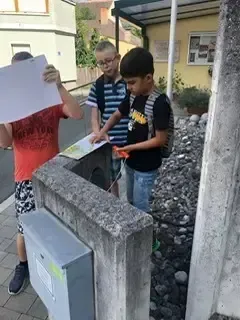 Drei Kinder, eines hält eine Karte, stehen neben einer Steinstruktur. Ein Junge trägt einen Rucksack und ein anderer hat ein rotes Shirt an.