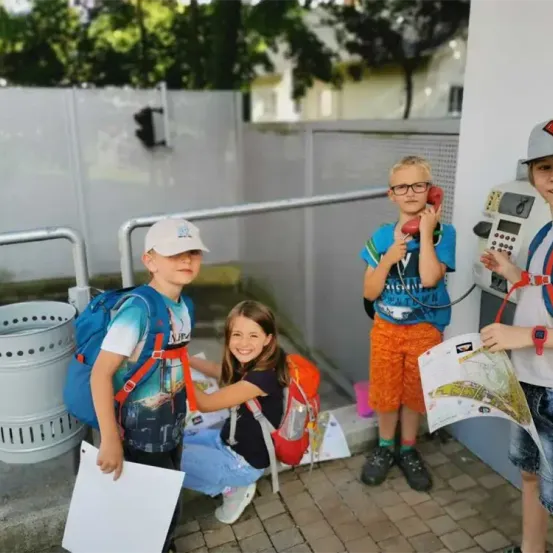 Vier Kinder stehen draußen in der Nähe einer Telefonzelle. Sie tragen Rucksäcke und halten Karten und Papiere. Ein Kind spricht in ein rotes Telefon. Ein Mülleimer steht in der Nähe.