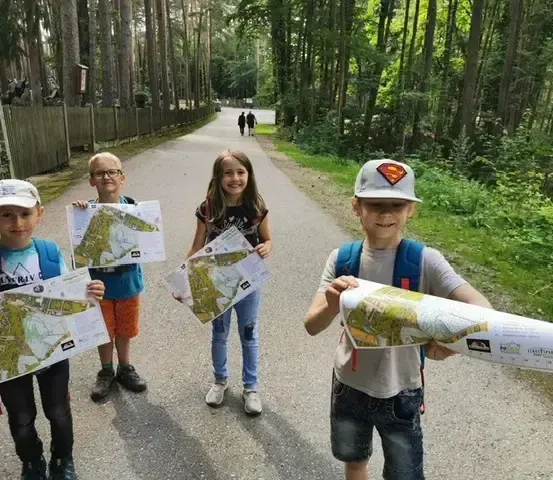 Vier Kinder stehen auf einem Pfad, halten Karten in der Hand, und zwei weitere gehen in der Ferne. Sie sind für Outdoor-Aktivitäten gekleidet und scheinen sich auf einem Waldabenteuer zu befinden.