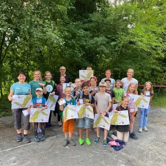 Eine Gruppe von Kindern und Erwachsenen posiert für ein Foto in einem Waldgebiet und hält Landkarten in der Hand.