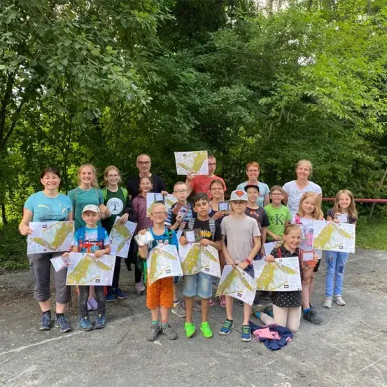 Eine Gruppe von Menschen, einschließlich Erwachsener und Kindern, steht draußen, hält Karten in der Hand und lächelt für ein Foto. Sie tragen Freizeitkleidung. Bäume und ein roter Zaun sind im Hintergrund zu sehen.