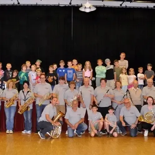 Eine Gruppe von Musikern, einschließlich Kinder, steht auf einer Bühne mit Musikinstrumenten. Sie sind in grauen Hemden und Jeans gekleidet. Die Bühne ist mit Holzboden bedeckt. Ein schwarzer Vorhang hängt an der Wand hinter ihnen.