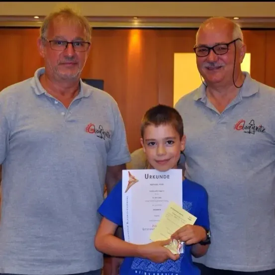 Drei Männer stehen neben einem jungen Jungen, der ein Zertifikat mit dem Text 'Urkunde' hält. Der Junge hält eine Medaille und das Zertifikat.