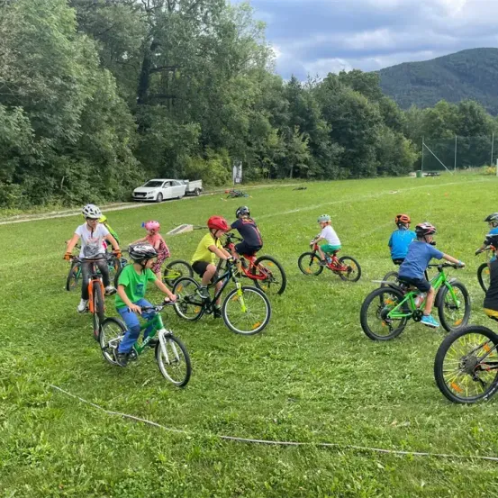 Kinder fahren auf einem Grasfeld mit einem Gebirgszug im Hintergrund Fahrräder. Sie tragen Helme und Turnschuhe. Ein Pickup-Truck und ein Auto sind auf der Straße geparkt.
