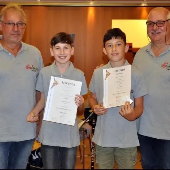 Zwei Jungen halten Urkunden in den Händen und stehen neben zwei älteren Männern. Die Jungen lächeln und halten Urkunden mit der Aufschrift Urkunde. Die Männer tragen Brillen und graue Poloshirts. Hinter ihnen ist ein unscharfer Hintergrund mit einem gelben Licht.