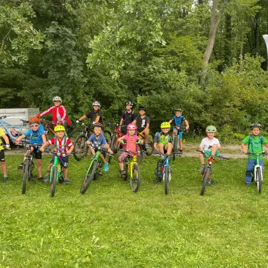Eine Gruppe junger Radfahrer in Helmen posiert für ein Foto auf einer Wiese, mit Fahrrädern und Bäumen im Hintergrund.